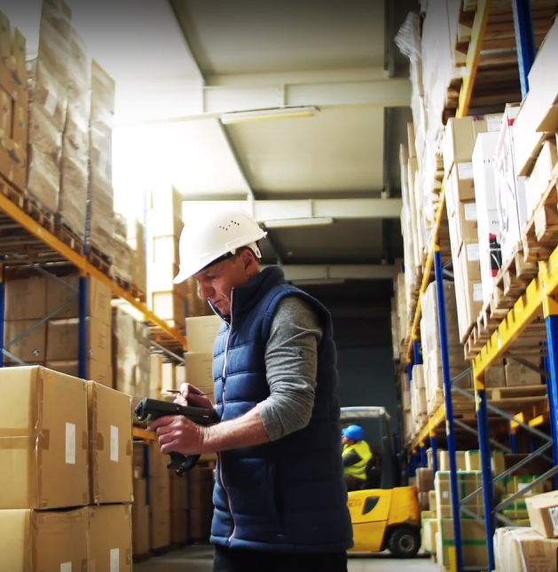 Warehouse worker in a hard hat using a handheld scanner to check boxes, with shelves of packages and a forklift in the background