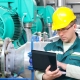 Engineer in safety helmet using a laptop to monitor and control industrial machinery in a manufacturing facility
