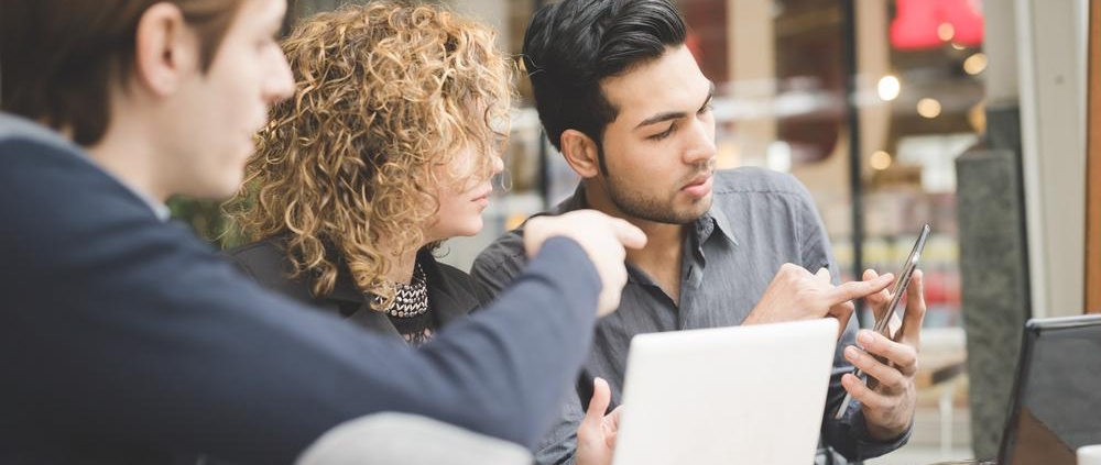 Three young professionals collaborating at a café table with laptops, tablet, and smartphone during a business meeting