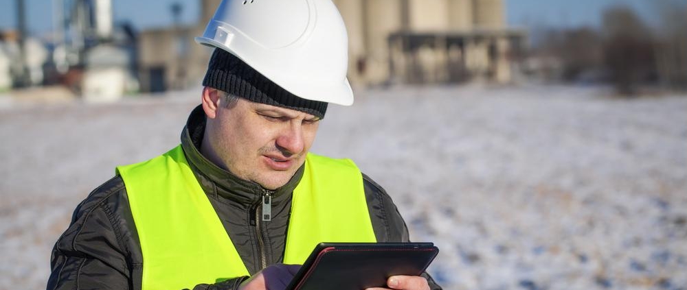 Construction worker in safety vest and helmet using a tablet outdoors at an industrial site