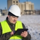Construction worker in safety vest and helmet using a tablet outdoors at an industrial site