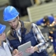 Two engineers wearing blue helmets using a tablet for work planning in a factory environment