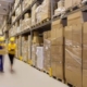 Warehouse interior with tall shelves stacked with cardboard boxes and two workers carrying a package