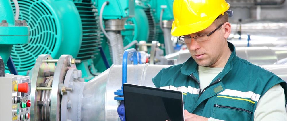 Engineer in safety helmet using a laptop to monitor and control industrial machinery in a manufacturing facility.