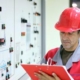 Engineer in a red safety helmet inspecting an electrical control panel and taking notes on a clipboard